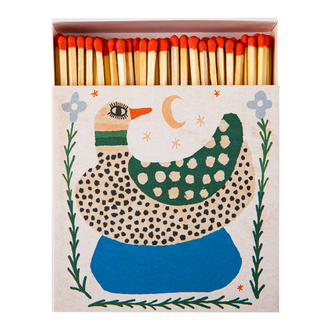 A decorative matchbox filled with wooden matches topped with red tips. The front of the box features a whimsical duck illustration in green, blue, black, and cream on a light beige background.