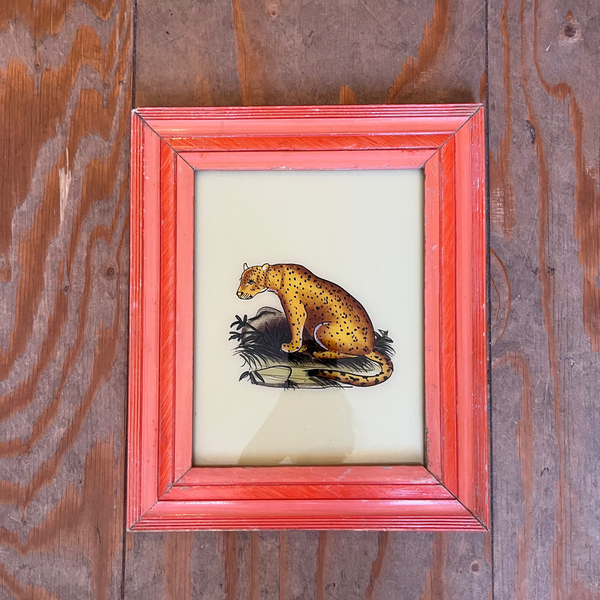 A framed vintage illustration of a leopard or cheetah sitting on a log, set in a reddish-orange frame against a rustic brown wooden plank background.