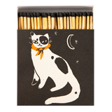 A matchbox-style match set with a dark charcoal cover featuring a white cat illustration with black patches and an orange bow tie, plus a crescent moon and stars. Wooden matches with black tips are visible at the top.
