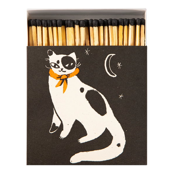 A matchbox-style match set with a dark charcoal cover featuring a white cat illustration with black patches and an orange bow tie, plus a crescent moon and stars. Wooden matches with black tips are visible at the top.