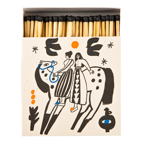 A matchbox filled with wooden matches, shown from above on a white background. The matchbox cover is cream-colored with a black line-art illustration of two riders on a spotted animal, accented with small orange and blue details.