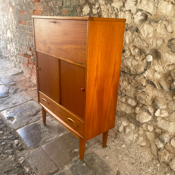 A mid-century modern wooden cabinet with a warm honey finish, featuring a top drawer, two lower doors, and a bottom drawer, set against a rustic stone wall.