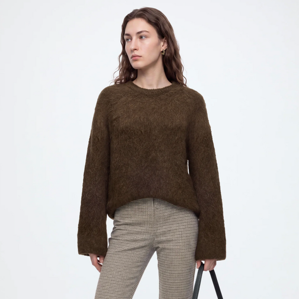 A model stands against a light gray studio background wearing a brown fuzzy long-sleeve sweater, beige plaid trousers, black loafers, and carrying a dark green shoulder bag.