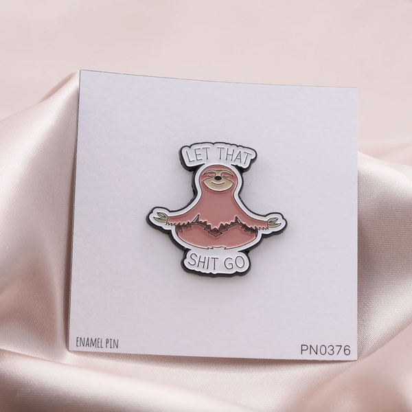A novelty enamel pin featuring a sloth meditating with the text 'LET THAT SHIT GO' on a white backing card, set against a draped light pink satin fabric background.