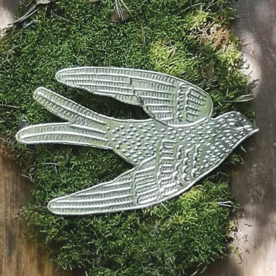A silver metal swallow ornament with textured detailing, displayed on a bed of green moss over a rustic wooden surface.