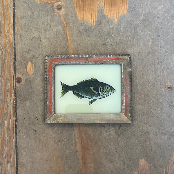 A small framed art piece featuring a painted fish on a pale background, mounted on a rustic wooden wall.