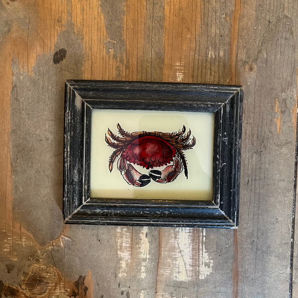 A small framed art piece featuring a red crab illustration on a cream background, set in a dark distressed frame and displayed on a rustic wooden surface.