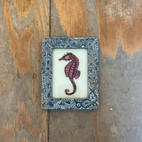 A small framed art print featuring a red seahorse on an off-white background, set in a blue and white patterned frame, displayed on a rustic wooden surface.