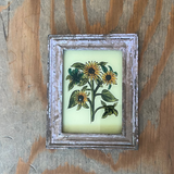 A small framed botanical art print featuring yellow sunflowers and green leaves on a pale yellow background, set in a distressed gray frame and placed on a rustic wooden surface.