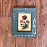 A small framed floral art print with a distressed teal-blue frame, featuring a red and orange flower illustration on a cream background, placed on a rustic wooden surface.