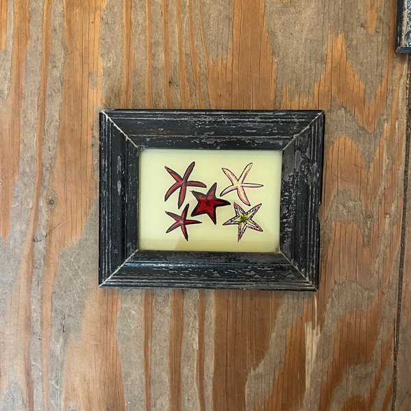 A small framed piece of wall art featuring three starfish in red, pink, and green on a cream background, mounted on a rustic wooden wall.