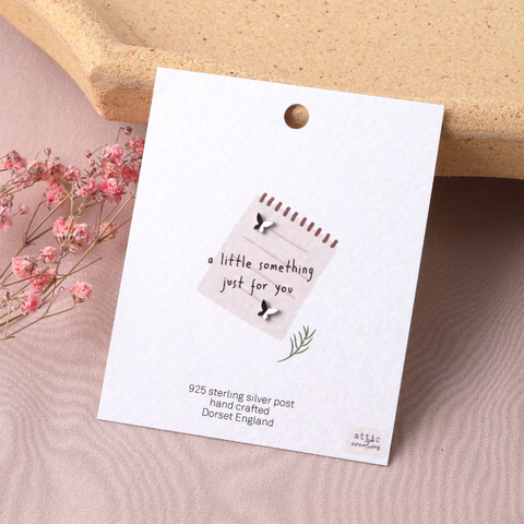 A sterling silver post pendant on a white display card, styled on a textured pink surface with dried pink flowers and a beige stone prop.
