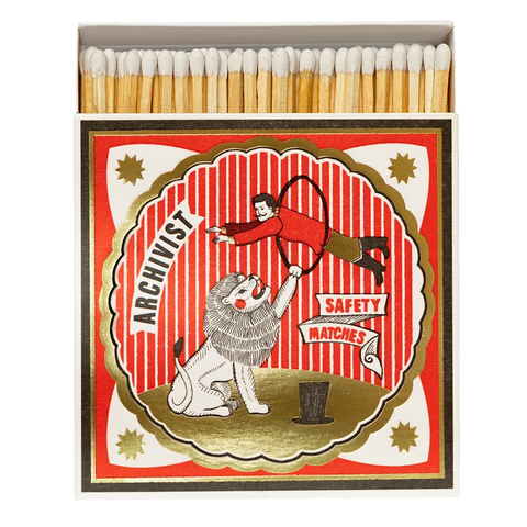 A vintage-style matchbox with wooden matches visible at the top and a illustrated front label reading 'ARCHIVIST' and 'SAFETY MATCHES.' The label shows a red-and-white striped circus scene with a lion and a person in a red outfit.