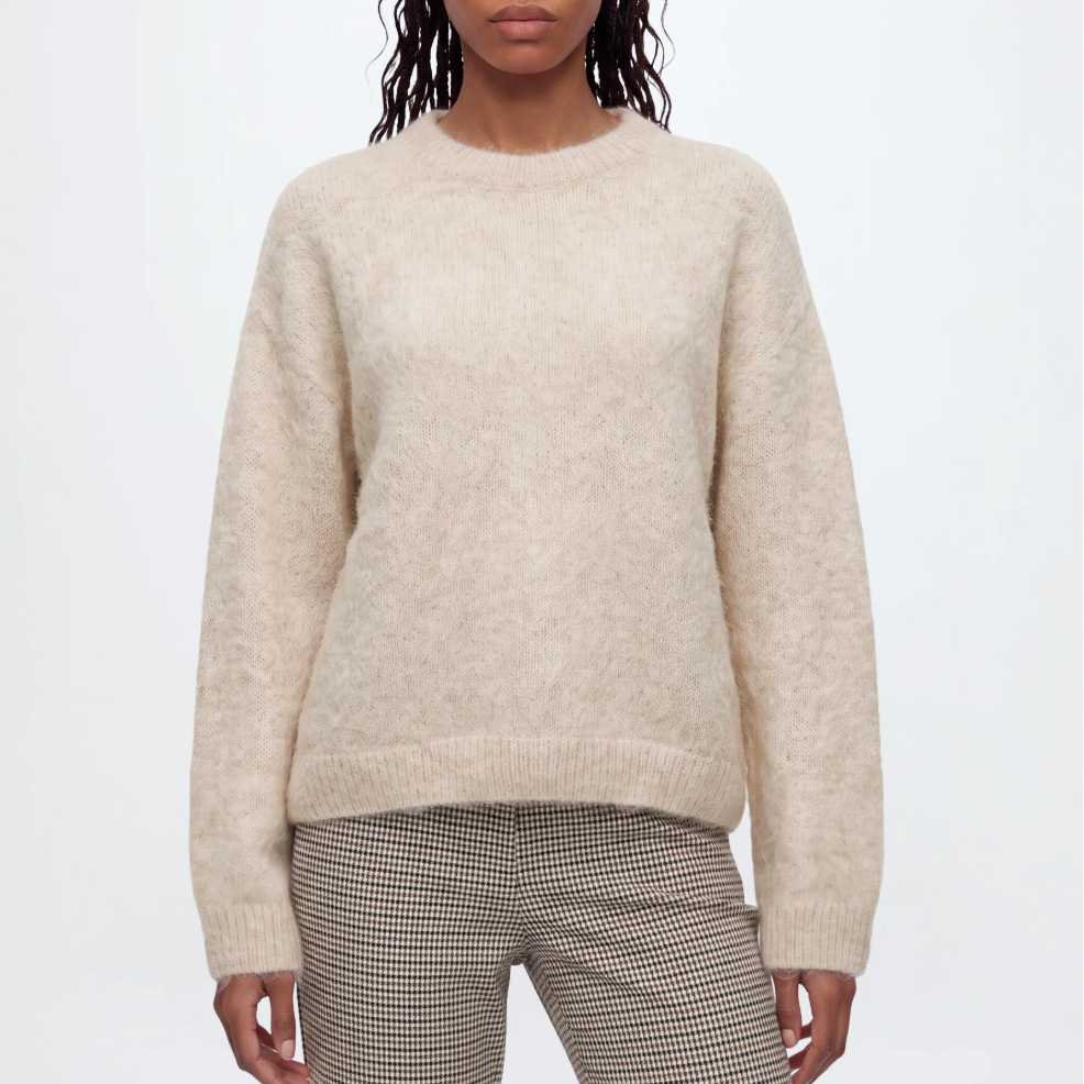 A woman modeling a cream-colored, long-sleeve alpaca knit sweater against a plain light gray background.
