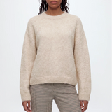 A woman modeling a cream-colored, long-sleeve alpaca knit sweater against a plain light gray background.