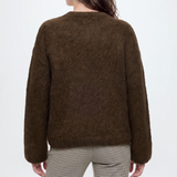 A woman shown from the back wearing an alpaca wool brown long-sleeve sweater and beige-and-black checkered trousers, against a plain light gray studio background.
