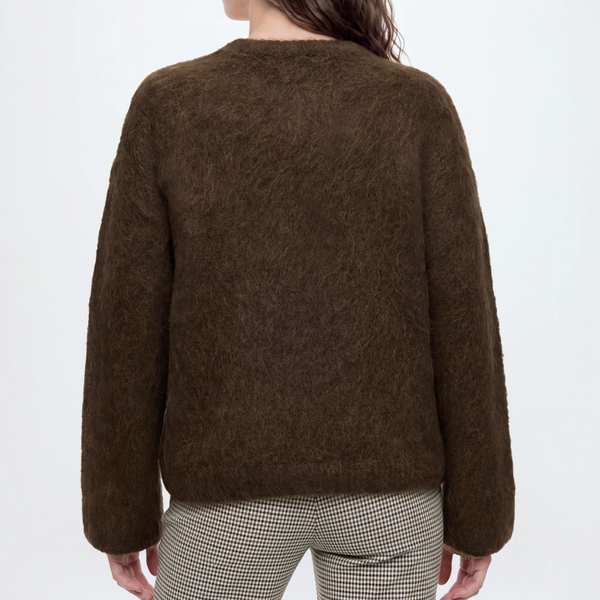 A woman shown from the back wearing an alpaca wool brown long-sleeve sweater and beige-and-black checkered trousers, against a plain light gray studio background.