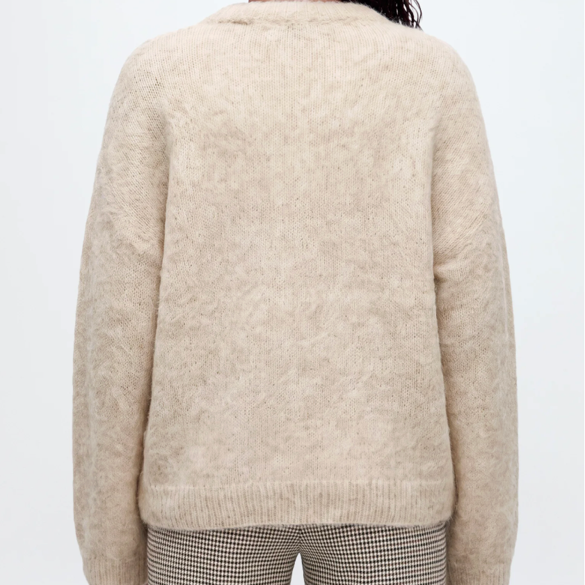 A woman viewed from behind wearing a cream-colored, fuzzy alpaca knit sweater and striped pants against a light gray studio background.
