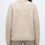 A woman viewed from behind wearing a cream-colored, fuzzy alpaca knit sweater and striped pants against a light gray studio background.