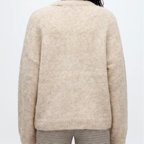A woman viewed from behind wearing a cream-colored, fuzzy alpaca knit sweater and striped pants against a light gray studio background.