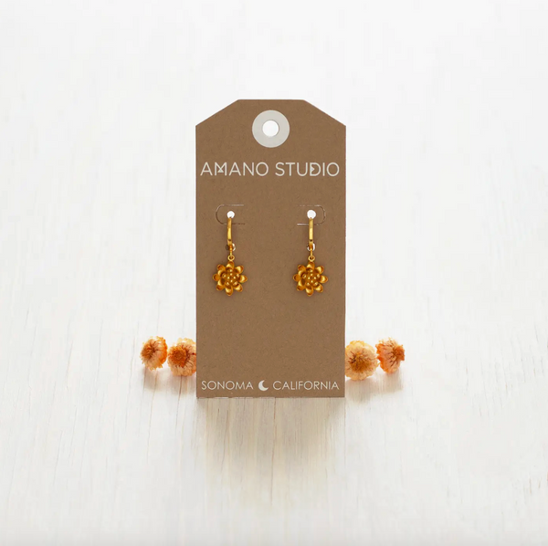 Gold earrings on a 'Amano Studio' branded card with dried flowers on a white background