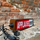 A vintage-style rectangular sign with a wooden frame and a red front panel reading 'APPLAUSE' in large white letters, with a black power cord attached, photographed against a brick wall and stone floor.