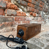 A wooden rectangular power adapter or charger with a black plug, shown resting on a stone surface against a red brick wall. The black cord and plug are visible in the foreground.