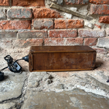 A vintage wooden chest or box with a power cord attached, resting on a stone floor against a red brick wall.