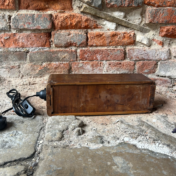 A vintage wooden chest or box with a power cord attached, resting on a stone floor against a red brick wall.