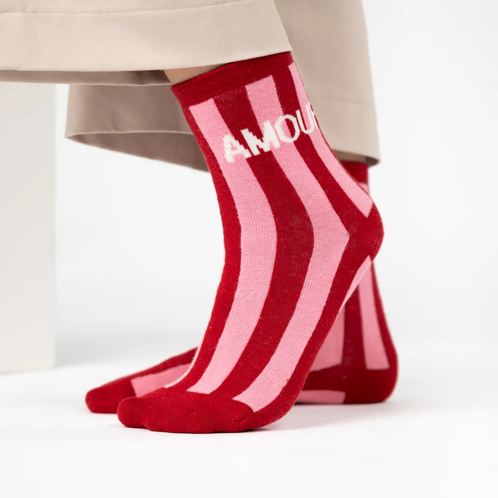 Red and pink striped ankle socks with the word 'AMOUR' in white on the side, shown on a model's feet with beige trousers visible at the top.