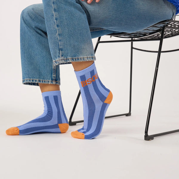 A person seated on a black wire chair wearing blue striped crew socks with orange toes and heels, paired with light blue jeans, against a clean white studio background.