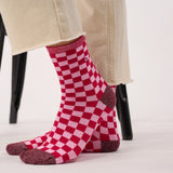 A pair of red and pink checkerboard crew socks worn on feet, with beige pants visible above the ankles and a black chair leg in the background.