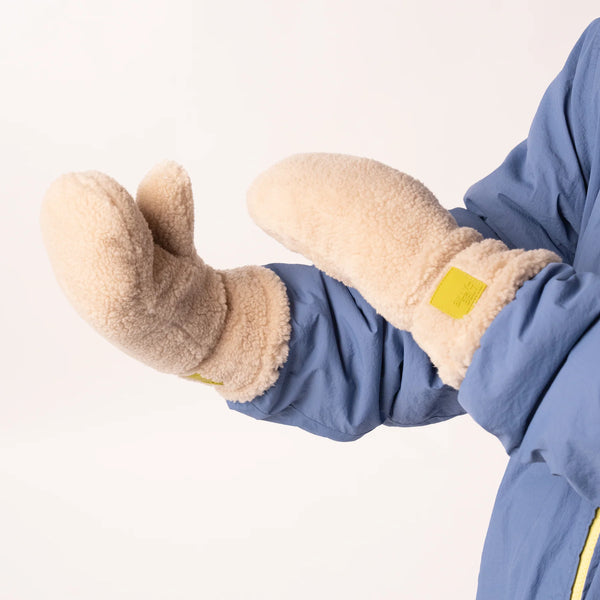 A close-up of a person wearing beige fleece mittens with a blue jacket against a plain white background.