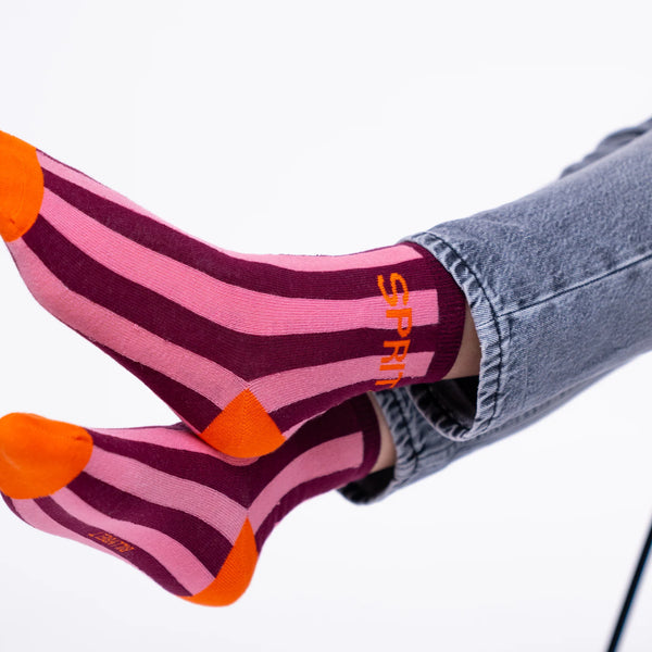 Pink and burgundy striped socks with bright orange toes and heels, worn with gray jeans. The word 'SPRING' is printed vertically in orange on the side of one sock.