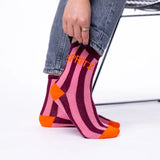 A person wearing pink and dark purple striped socks with bright orange toes and heels, with the word 'SPRITZ' embroidered in orange on the side. The person is wearing blue jeans and sitting on a white chair.