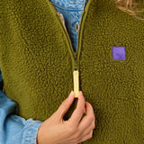 Close-up of a person wearing an olive green sherpa fleece zip-up vest over a light blue denim shirt, with a hand holding the zipper pull.