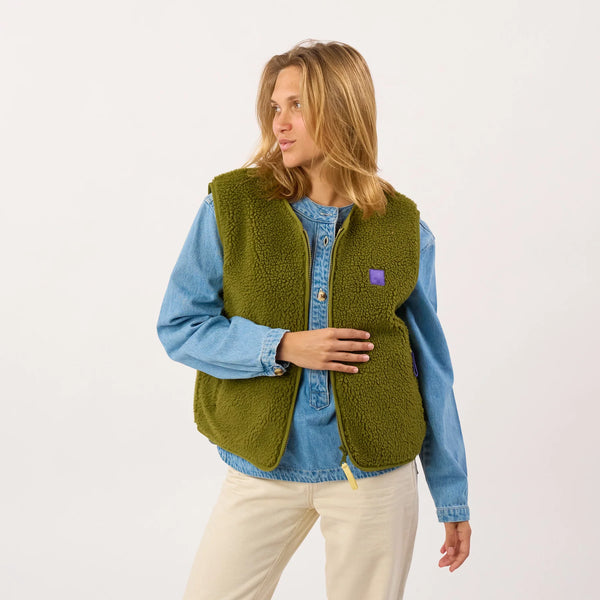 A woman wearing a sage green sherpa-lined zippered vest over a light blue denim shirt and cream pants, photographed against a clean white studio background.