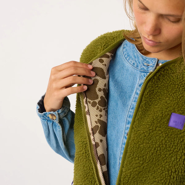 A close-up of a person wearing an olive green sherpa fleece zip jacket over a light blue denim shirt, showing the interior pattern lining and a purple brand patch on the chest.