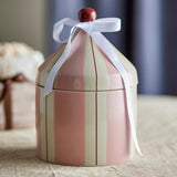 A round ceramic lidded container with vertical pink and cream stripes, decorated with a white ribbon bow and a small red top knob, set on a beige fabric surface with a blurred background.