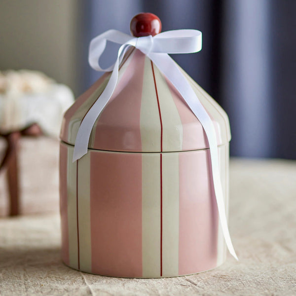 A round ceramic lidded container with vertical pink and cream stripes, decorated with a white ribbon bow and a small red top knob, set on a beige fabric surface with a blurred background.