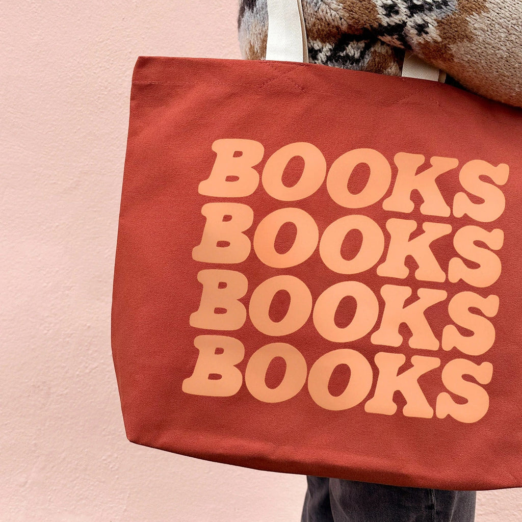 A terracotta red canvas tote bag with the word 'BOOKS' printed in a large peach-colored retro font, repeated vertically to read 'BOOKS BOOKS BOOKS BOOKS'. The bag is carried over the shoulder of a person wearing a brown, beige, and black patterned knit sweater against a soft pink wall.