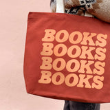A terracotta red canvas tote bag with the word 'BOOKS' printed in a large peach-colored retro font, repeated vertically to read 'BOOKS BOOKS BOOKS BOOKS'. The bag is carried over the shoulder of a person wearing a brown, beige, and black patterned knit sweater against a soft pink wall.