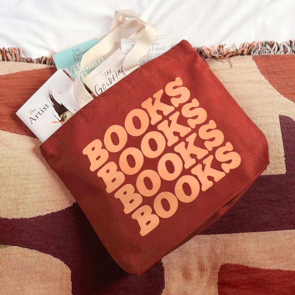 A terracotta red canvas tote bag with the word 'BOOKS' printed repeatedly in peach-colored letters, filled with books and resting on a patterned rug with a white blanket behind it.