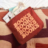 A terracotta red canvas tote bag with the word 'BOOKS' printed repeatedly in peach-colored letters, filled with books and resting on a patterned rug with a white blanket behind it.