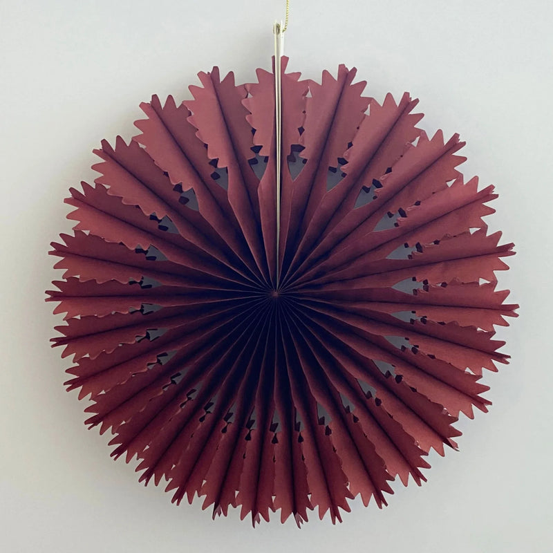 A circular pleated paper fan decoration in a deep burgundy red color, hanging against a plain off-white wall.
