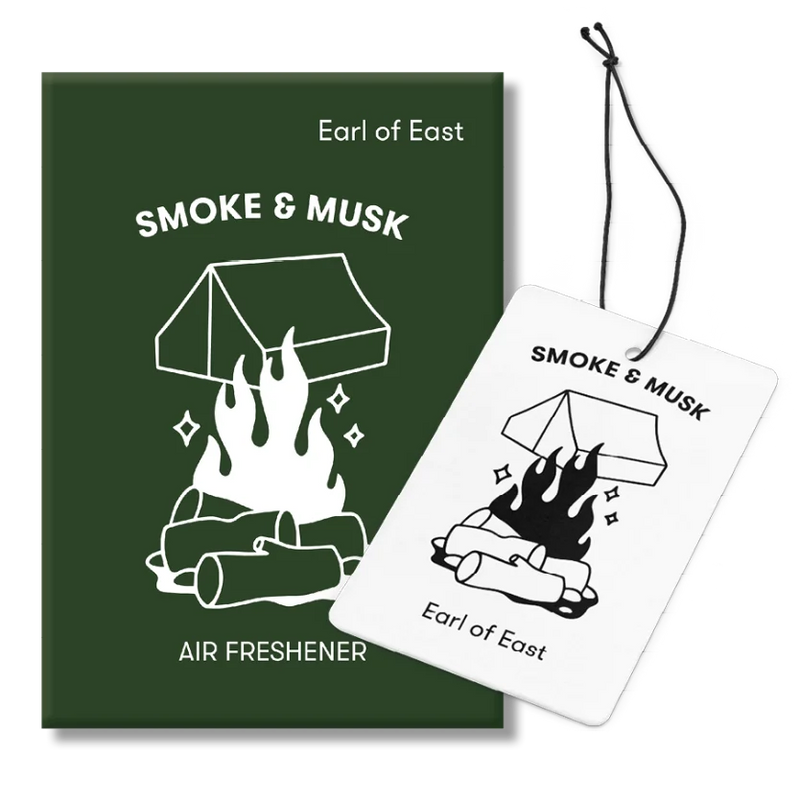 Earl of East Smoke & Musk air freshener packaging, featuring a dark green rectangular box with a white campfire and tent illustration, alongside a white rectangular car air freshener card with a black hanging string.