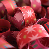 Close-up of red and pink patterned gift wrapping paper rolls with white starburst designs and small gold and teal accents, set against a softly blurred holiday background.