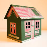 A handcrafted paper house model with a dark green body, pink and white striped roof, and orange window frames, set against a warm beige background.