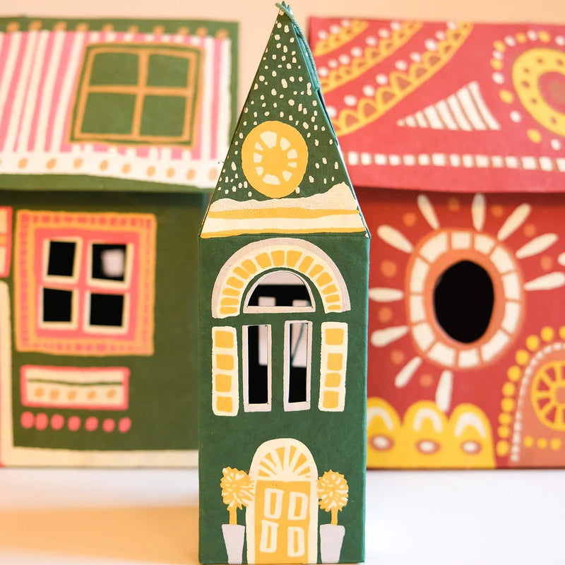 A small hand-painted house-shaped ornament or toy stands in the center, with two larger matching decorative houses blurred in the background against a warm beige wall and white surface.