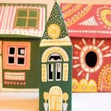 A small hand-painted house-shaped ornament or toy stands in the center, with two larger matching decorative houses blurred in the background against a warm beige wall and white surface.